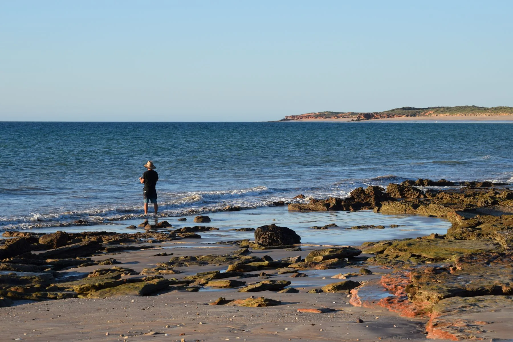 An angler beside clear blue water on a rocky beach